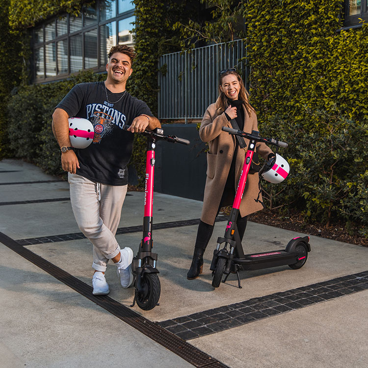 Promotional image for AA Member Benefit and Flamingo e-scooters. Two people stand outdoors beside bright pink-and-black electric scooters on a paved walkway next to a modern building covered in green climbing plants. Each person is holding a white helmet with a pink stripe. The AA Member Benefit logo appears in the top-left corner, featuring a yellow circle with “AA” and the text “Member Benefit.”