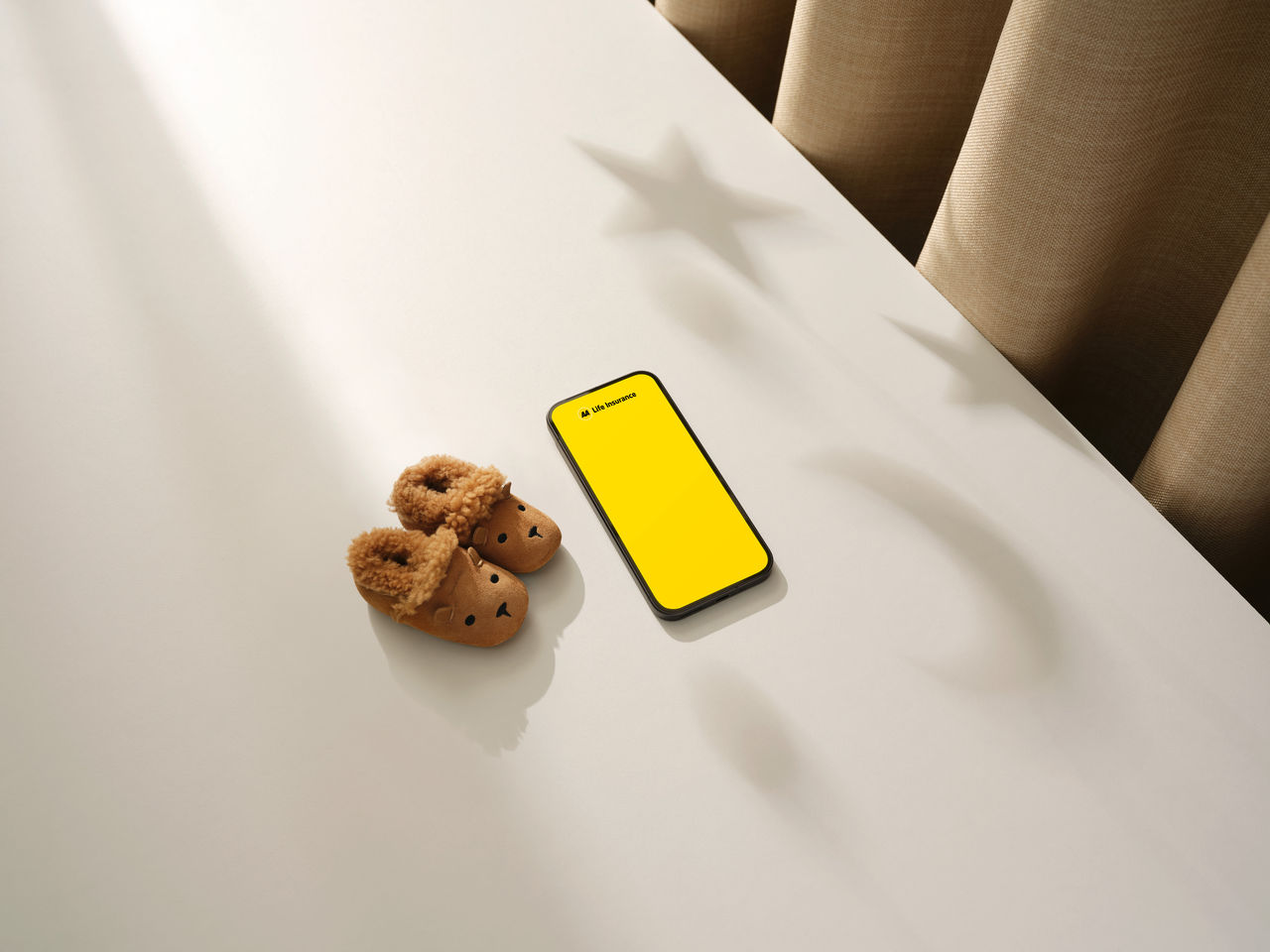 A smartphone displaying a bright yellow life insurance app screen sits on a white tabletop beside a pair of brown fleece-lined children's slippers shaped like animal faces. Warm sunlight streams across the surface, casting soft shadows, including star-shaped silhouettes from an unseen hanging decoration. Beige curtains in the background create a cozy, home-like atmosphere with ample negative space around the objects.