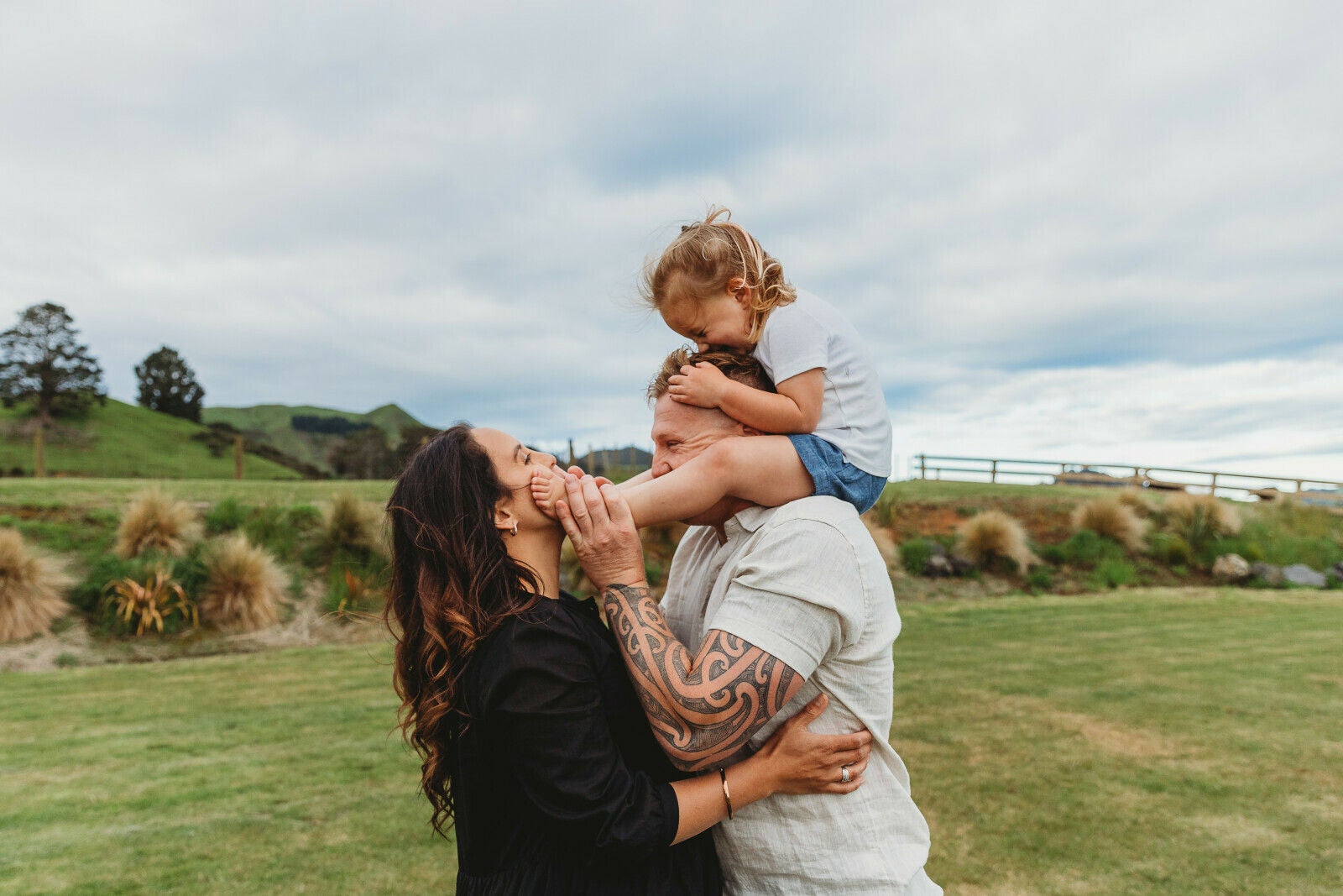 An adult wearing a light shirt stands outdoors in a grassy rural setting, holding a young child on their shoulders. Another adult with long dark hair reaches up toward the child, gently holding the child’s foot. The group is positioned on a green lawn with rolling hills, bushes, and a wooden fence in the background under a cloudy sky.