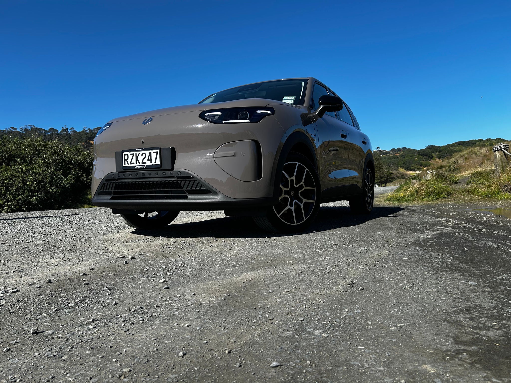 Low-angle front three-quarter view of a light-coloured Leapmotor B03X electric SUV parked on a gravel road. The image highlights the vehicle’s smooth front-end design, LED headlights, and multi-spoke alloy wheels. Coastal vegetation and sandy dunes line the roadside, while a clear blue sky provides a bright backdrop. Strong sunlight casts defined shadows beneath the vehicle, emphasizing its ground clearance and stance.