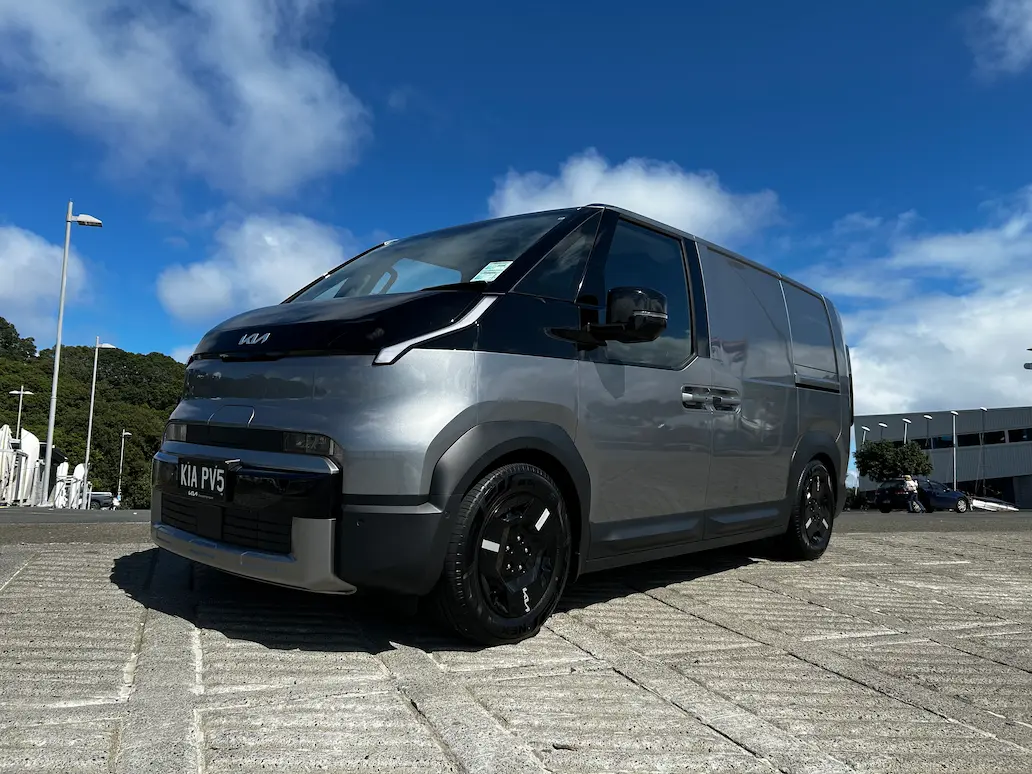 Front three-quarter view of a silver Kia PV5 electric cargo van parked outdoors under a bright blue sky.