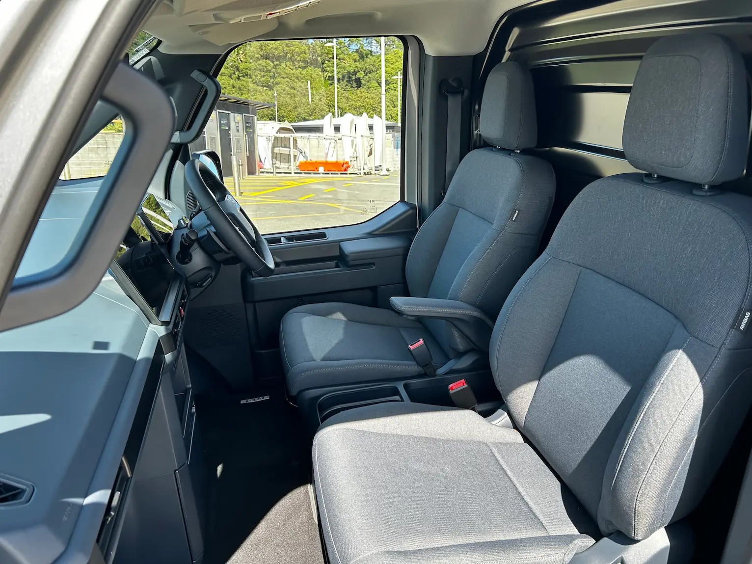 Front cabin of a Kia PV5 cargo van showing two grey fabric seats, a digital dashboard, and a bulkhead partition behind the seats.