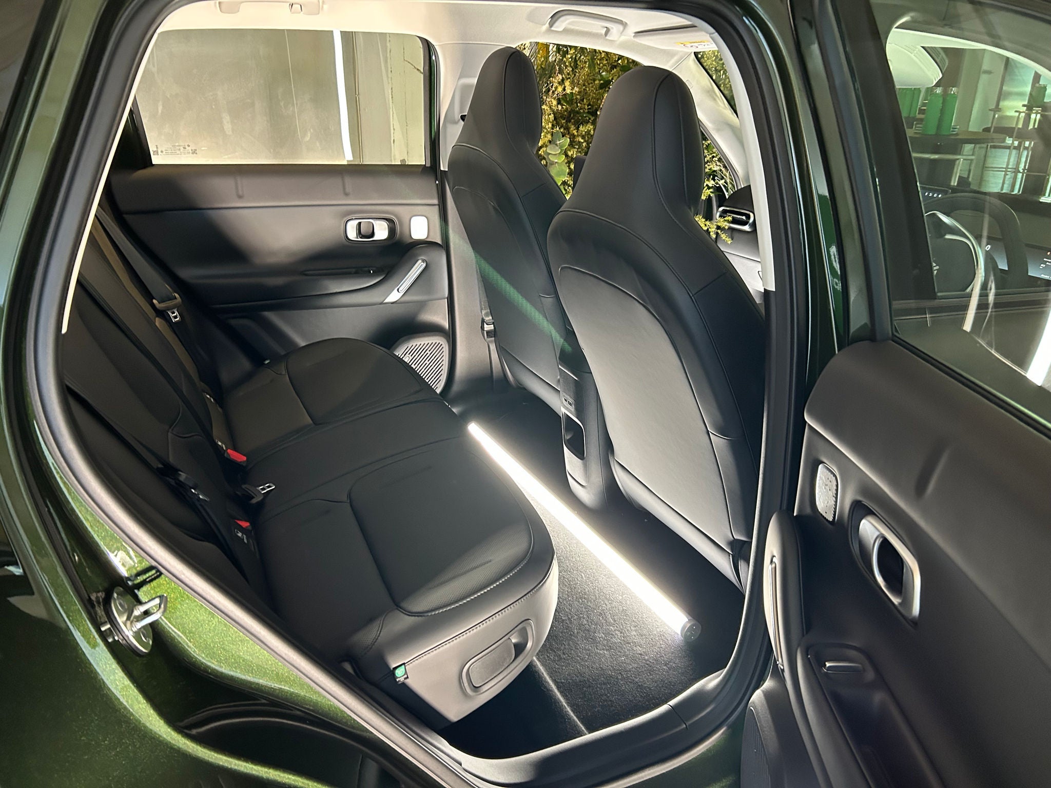 View into the rear passenger area of a dark green SUV with the rear door open. The interior features black upholstered seats, a flat rear bench, and two front seats with slim backs. A bright light placed on the floor illuminates the rear footwell, highlighting the legroom between the front and rear seats. The door panels include silver trim accents and integrated speakers, while greenery and event décor are visible through the windows in the background.