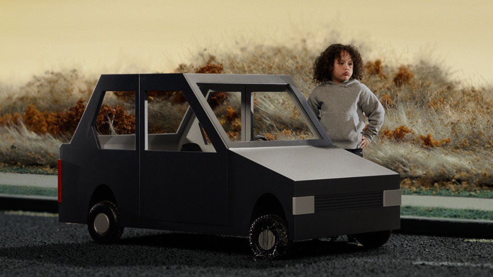 A person in a gray hooded sweatshirt stands beside a black, angular model car parked on a road. Dry grasses and shrubs line the background, while the dark road surface in the foreground appears wet and reflective.