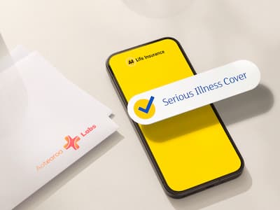 Top-down view of a minimalist tabletop scene featuring a smartphone with a bright yellow life insurance app screen and a floating "Serious Illness Cover" confirmation badge. Two white envelopes sit beside the phone, along with two clear glasses of water. Soft natural light casts gentle shadows across the light-colored surface, creating a clean and modern insurance-themed setting.