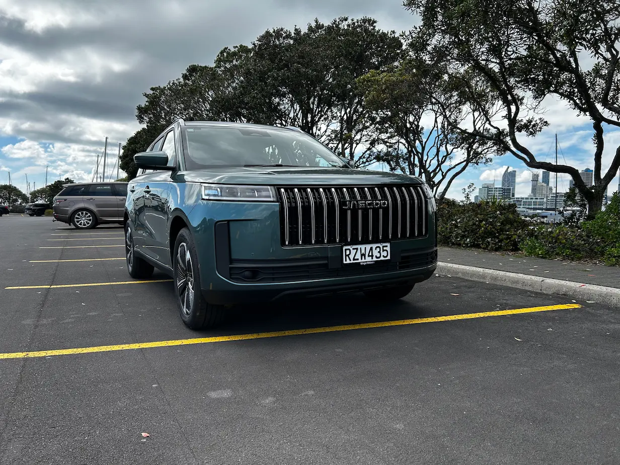 Dark green JAECOO SUV parked in a waterfront car park, viewed from the front with trees and Auckland's city skyline in the background under cloudy skies.