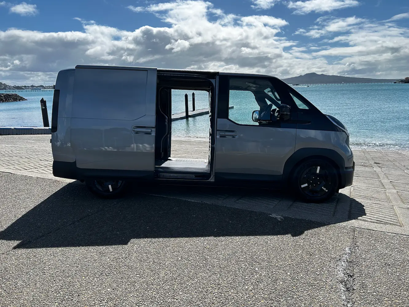 Side view of a Kia PV5 electric cargo van parked beside the water, with the sliding side door open to reveal the cargo area.