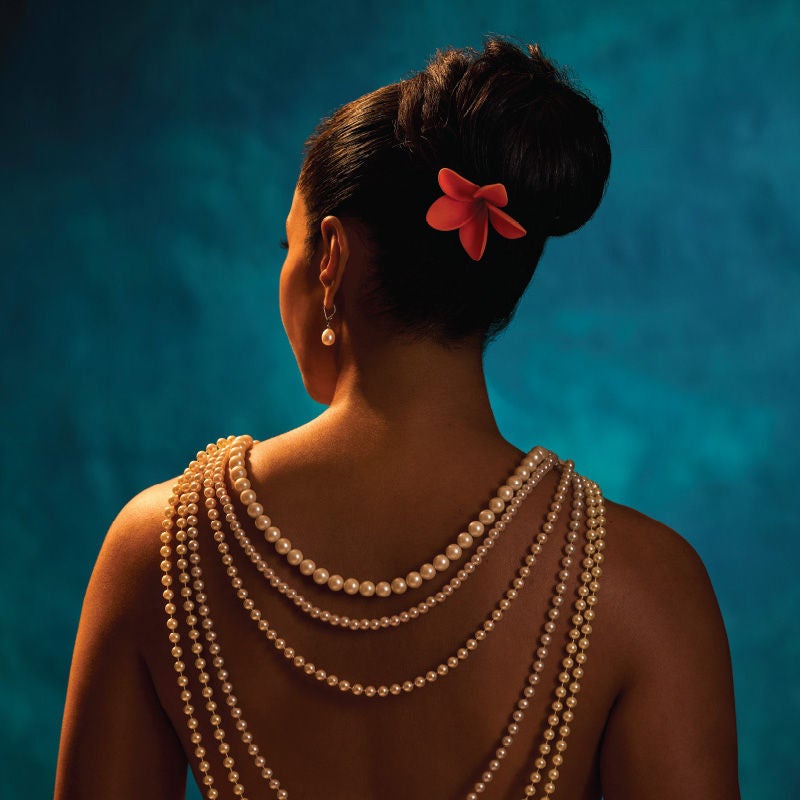 Person shown from behind with dark hair pinned up and decorated with a pink flower. Multiple strands of white pearls drape across the bare back and shoulders, with a pearl earring visible. The subject is illuminated by warm lighting against a textured blue background.