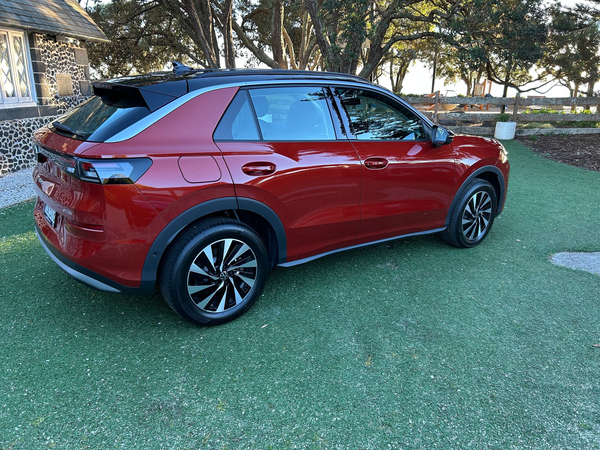 Rear three-quarter side view of a red Volkswagen T-Roc SUV parked on a grass-covered area beside a historic stone building. The vehicle features a contrasting black roof, black wheel arch cladding, tinted windows, roof spoiler, and multi-spoke alloy wheels. The sculpted bodywork, rear LED taillights, and sloping C-pillar design are visible from this angle. Mature trees, landscaped grounds, and timber fencing form the background, while natural daylight highlights the metallic red paint and exterior styling details. 