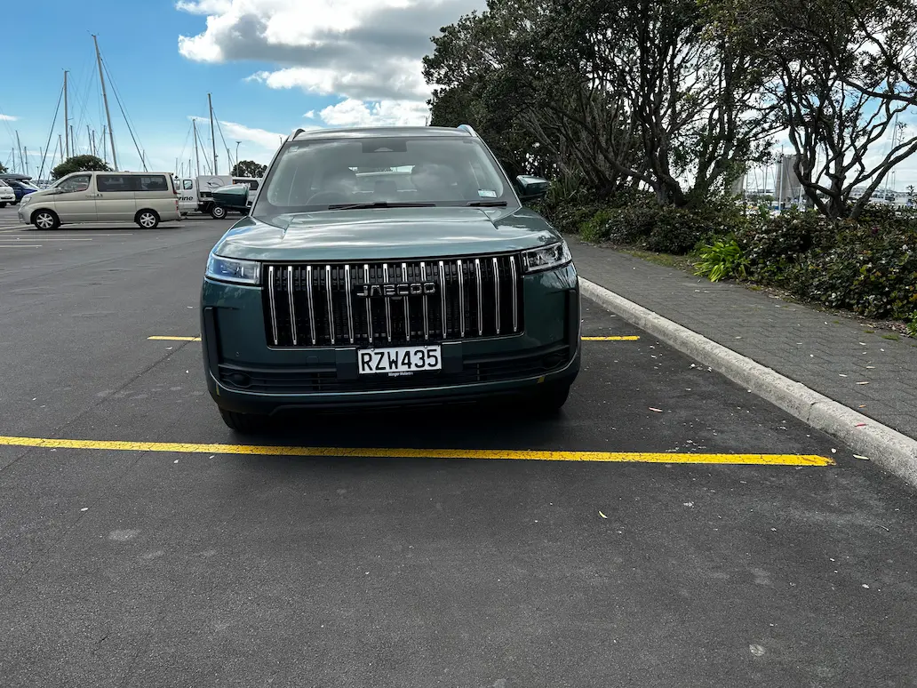 Dark green JAECOO SUV parked in a waterfront car park, viewed from the front with trees and Auckland's city skyline in the background under cloudy skies.