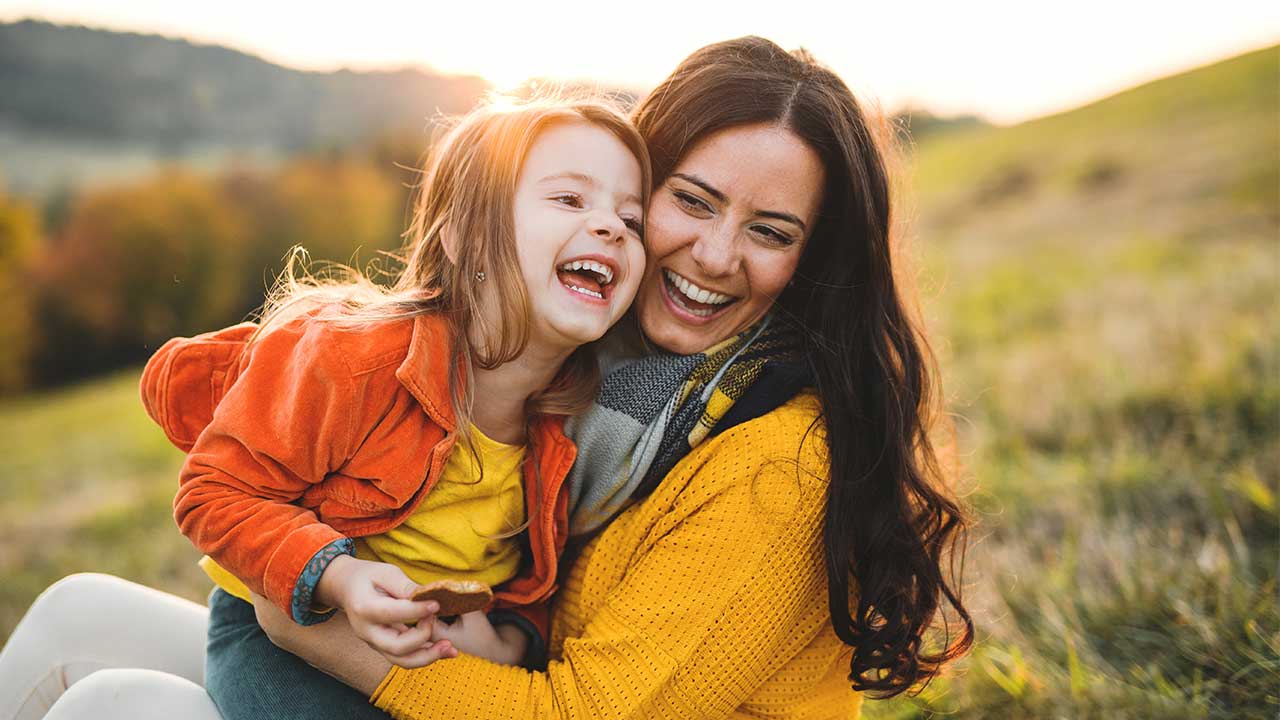 A mother and her young daughter laughing together on a hillside.