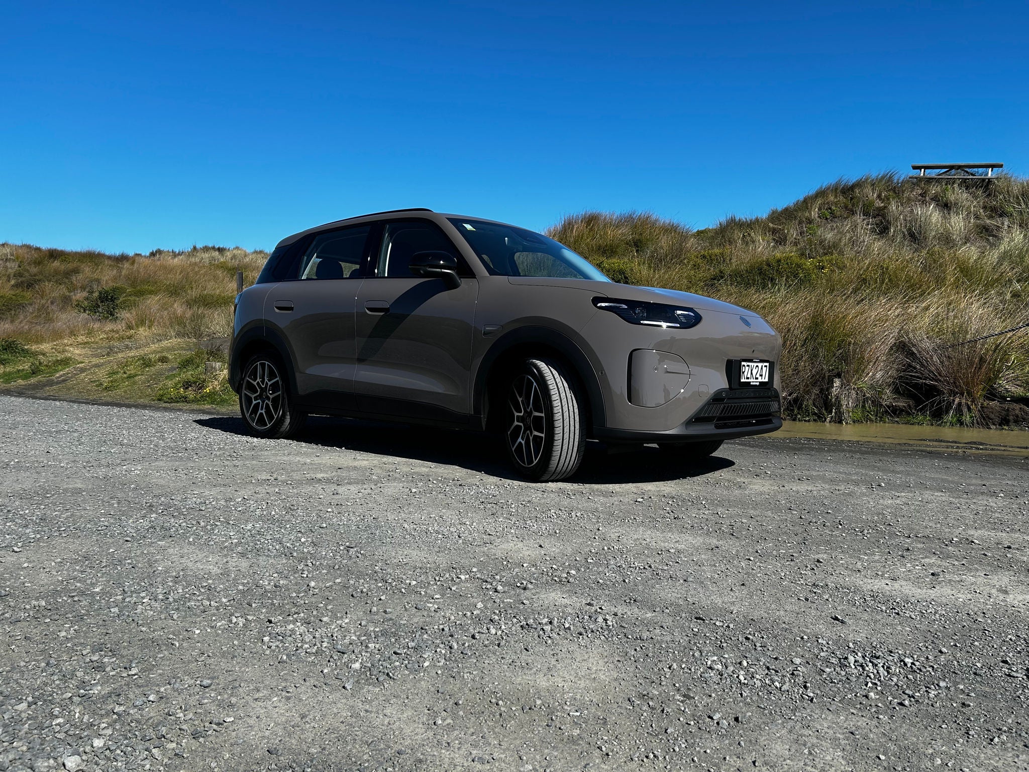 Light-colored SUV parked on a gravel road in a coastal dune landscape, photographed from the front three-quarter angle under a clear blue sky. Tall grass-covered sand dunes surround the vehicle, with bright sunlight casting strong shadows and highlighting the car's modern design. A vehicle license plate is visible on the front, and a small lookout platform appears on a dune in the background.