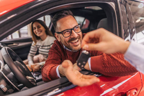 Smiling customer being handed car keys in the front seat of a red car, with a front-seat passenger smiling in the foreground