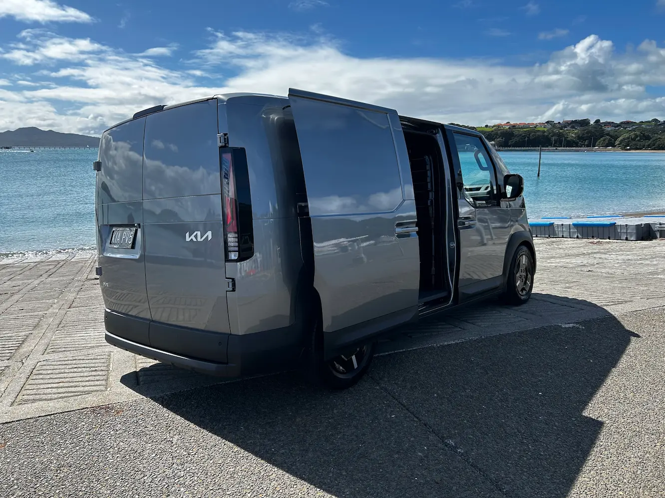Grey Kia PV5 electric van parked beside a waterfront boat ramp, with the side sliding door open and blue water and clouds visible in the background.