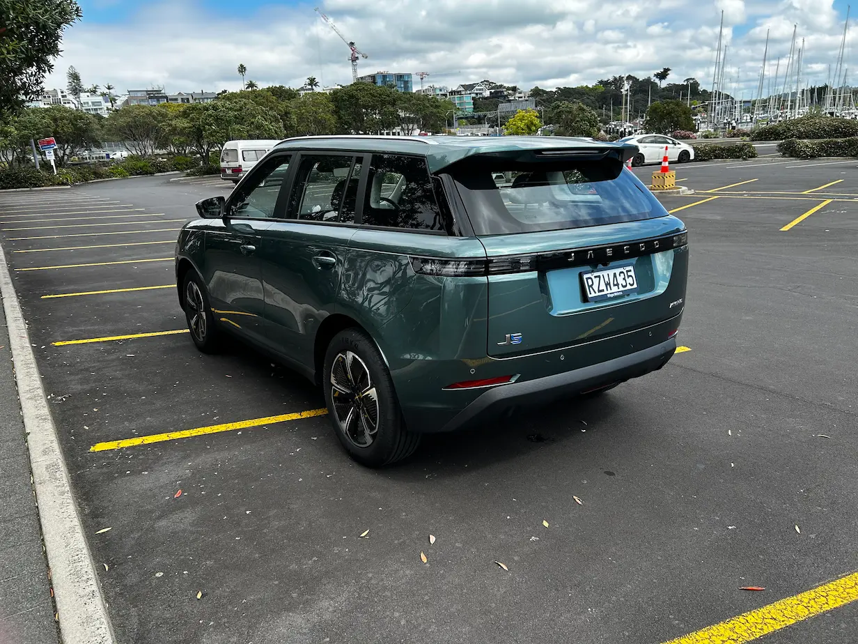 Rear three-quarter view of a dark green JAECOO J5 SUV parked in a waterfront car park, showing the tailgate, rear light bar, and alloy wheels.