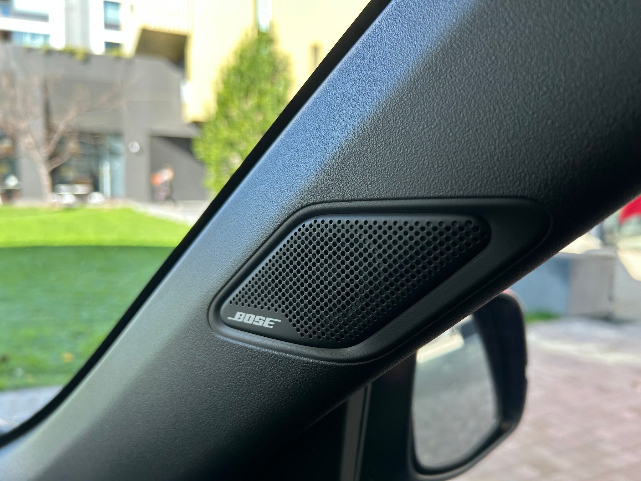 Close-up of a Bose-branded speaker mounted on the front A-pillar inside a Honda electric city car. The black perforated speaker grille is integrated into the textured interior trim beside the windscreen and side window. Part of the exterior side mirror is visible below the speaker, while a blurred background outside the vehicle shows a green lawn, paving, and modern buildings in daylight. The image highlights the vehicle's premium audio system and interior finish.