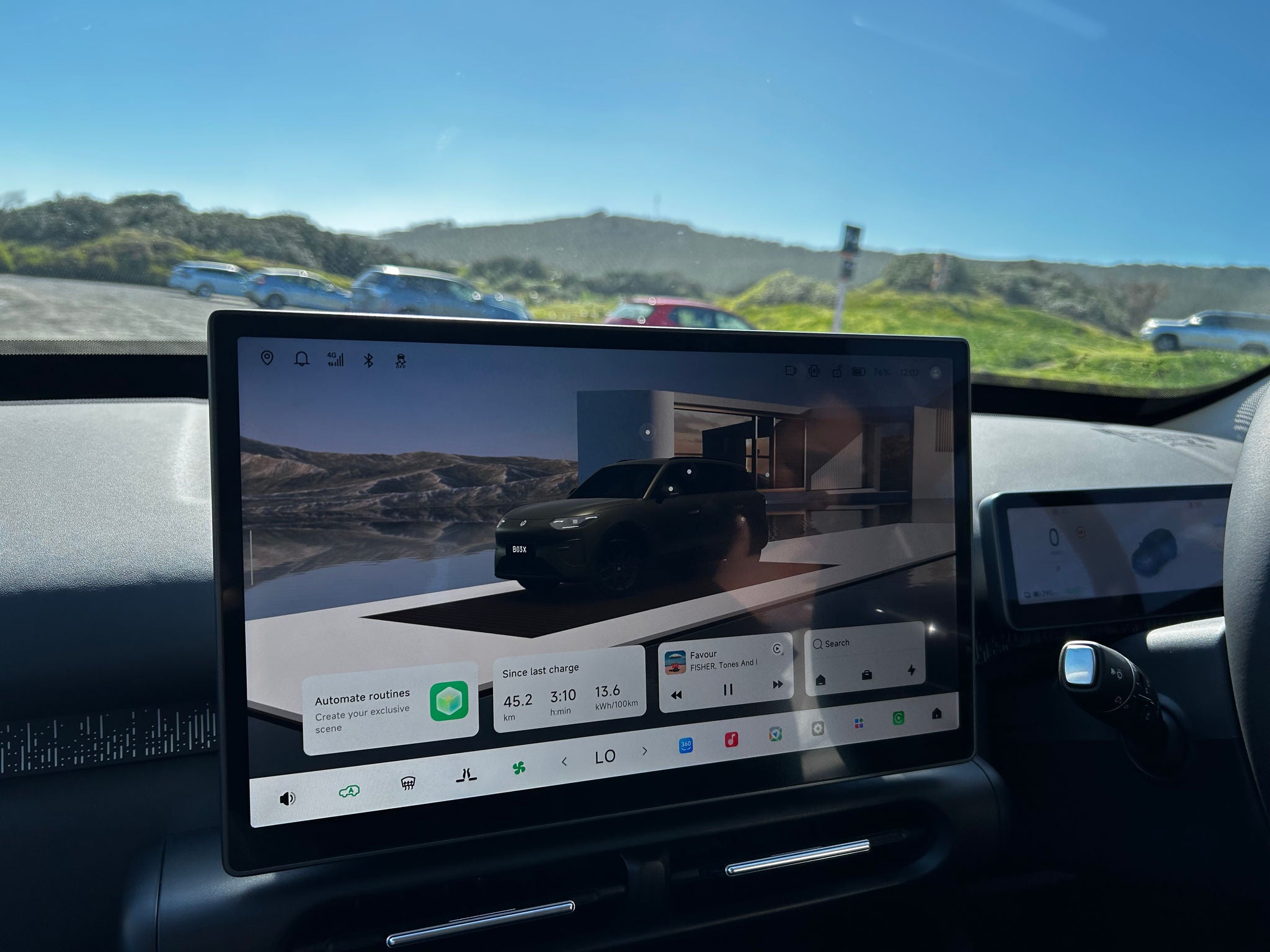 Close-up view of the Leapmotor B03X dashboard and infotainment system from the front passenger area. A large central touchscreen displays the vehicle interface, including charging data, media controls, climate settings and a 3D vehicle graphic. The right-hand-drive cockpit features a compact digital instrument cluster behind the steering wheel and slim air vents beneath the screen. Through the windscreen, a sunny outdoor scene with parked cars, grassy dunes and a clear blue sky is visible.