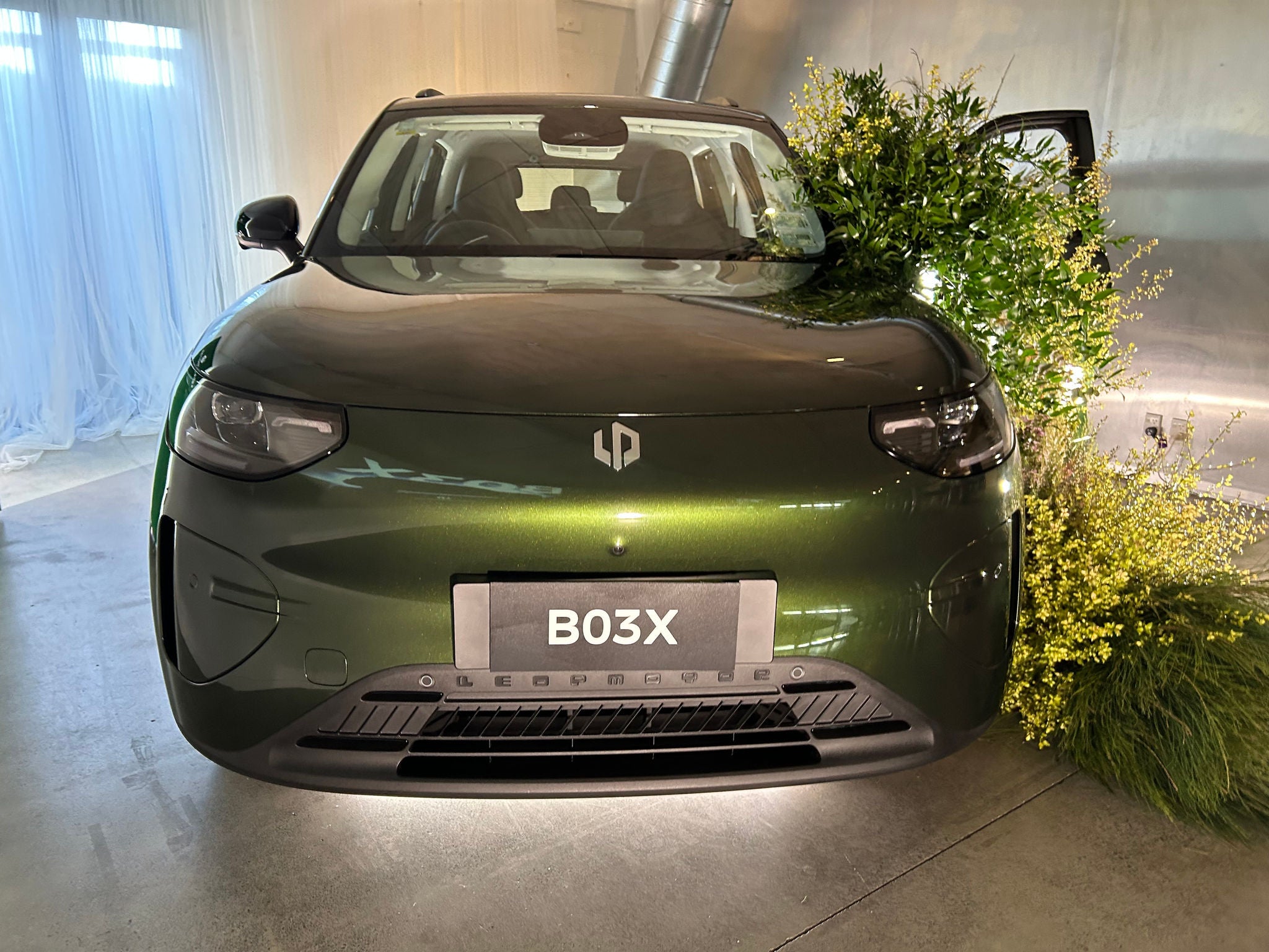 Front view of a dark green compact SUV displayed indoors at a vehicle launch or showroom event. The car is illuminated by spotlights and positioned on a polished concrete floor, with decorative green foliage and yellow flowering plants arranged around the front passenger-side corner. White draped curtains line the wall on the left, and a large ventilation duct is visible in the background. Visible text includes a front display plate reading “B03X” and branding across the lower front grille.