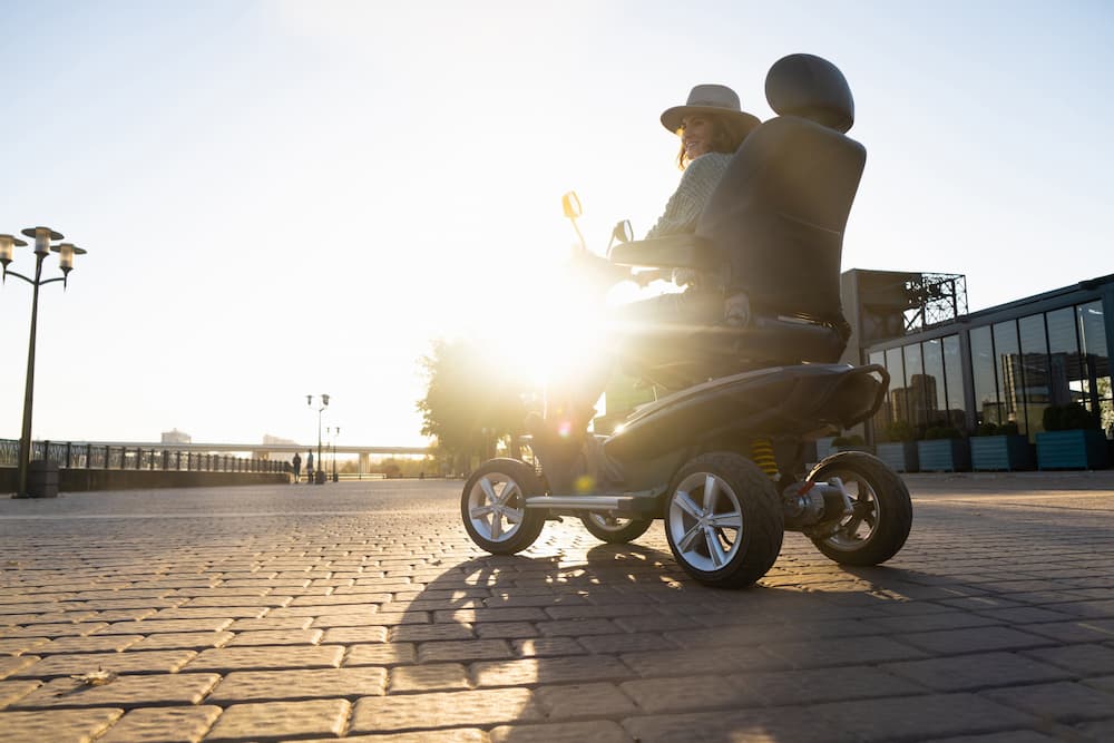 A person driving a large mobility scooter across a cobblestone plaza near a waterfront promenade. The scene is backlit by bright sunlight, creating lens flare and long shadows. Modern buildings, street lamps, and distant trees line the open public space, while the mobility scooter is shown prominently in the foreground with large wheels and a high-backed seat.