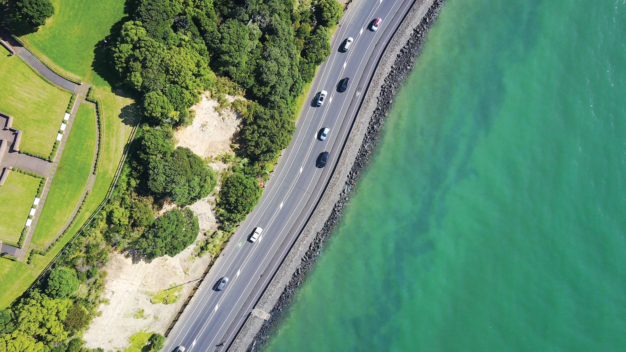 Top-down aerial view of a coastal roadway running parallel to a turquoise ocean. Several cars are traveling along the road, which is bordered by a rocky shoreline on one side and lush green trees, grassy parkland, and walking paths on the other.