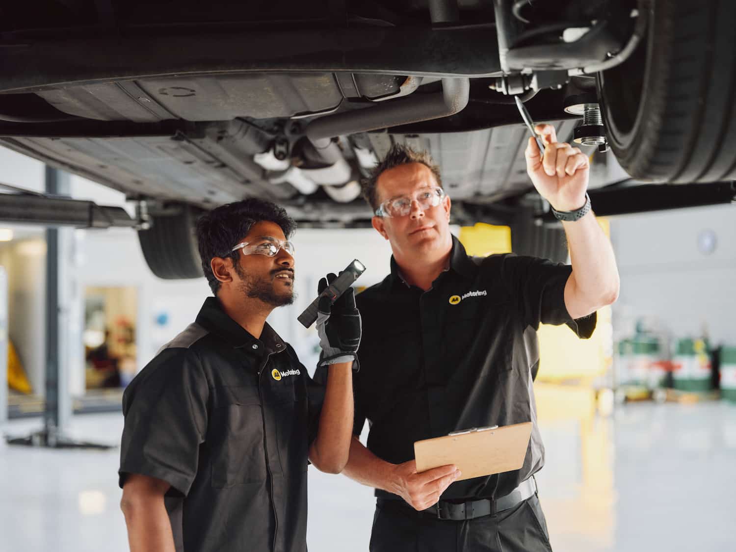 Two automotive technicians wearing safety glasses inspect the underside of a vehicle raised on a service hoist. One technician shines a flashlight toward vehicle components while the other points to an area near a wheel and holds a clipboard in a well-lit workshop.