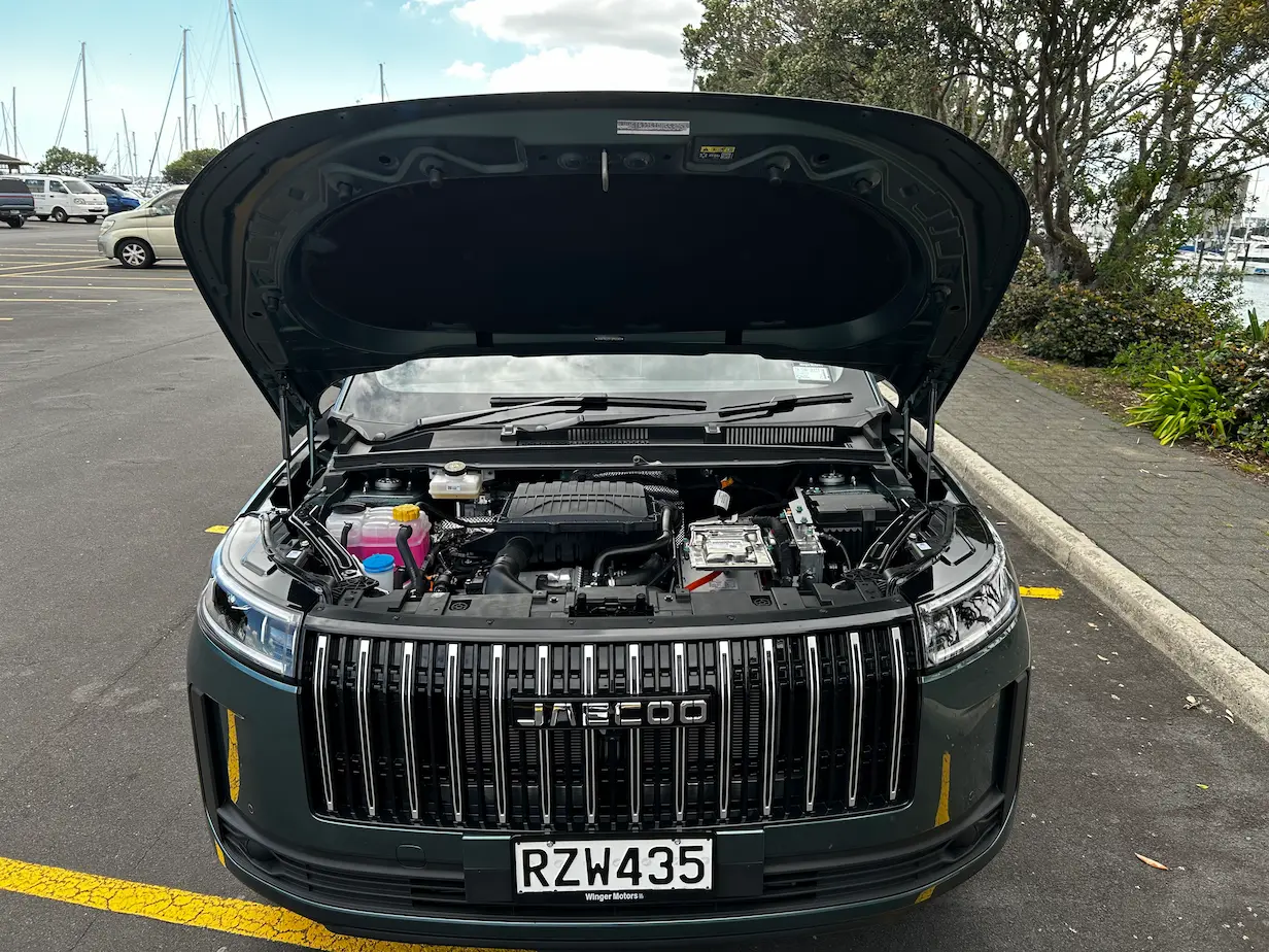 Front view of a dark green JAECOO SUV with the bonnet raised, showing the engine bay and grille while parked in a waterfront car park.