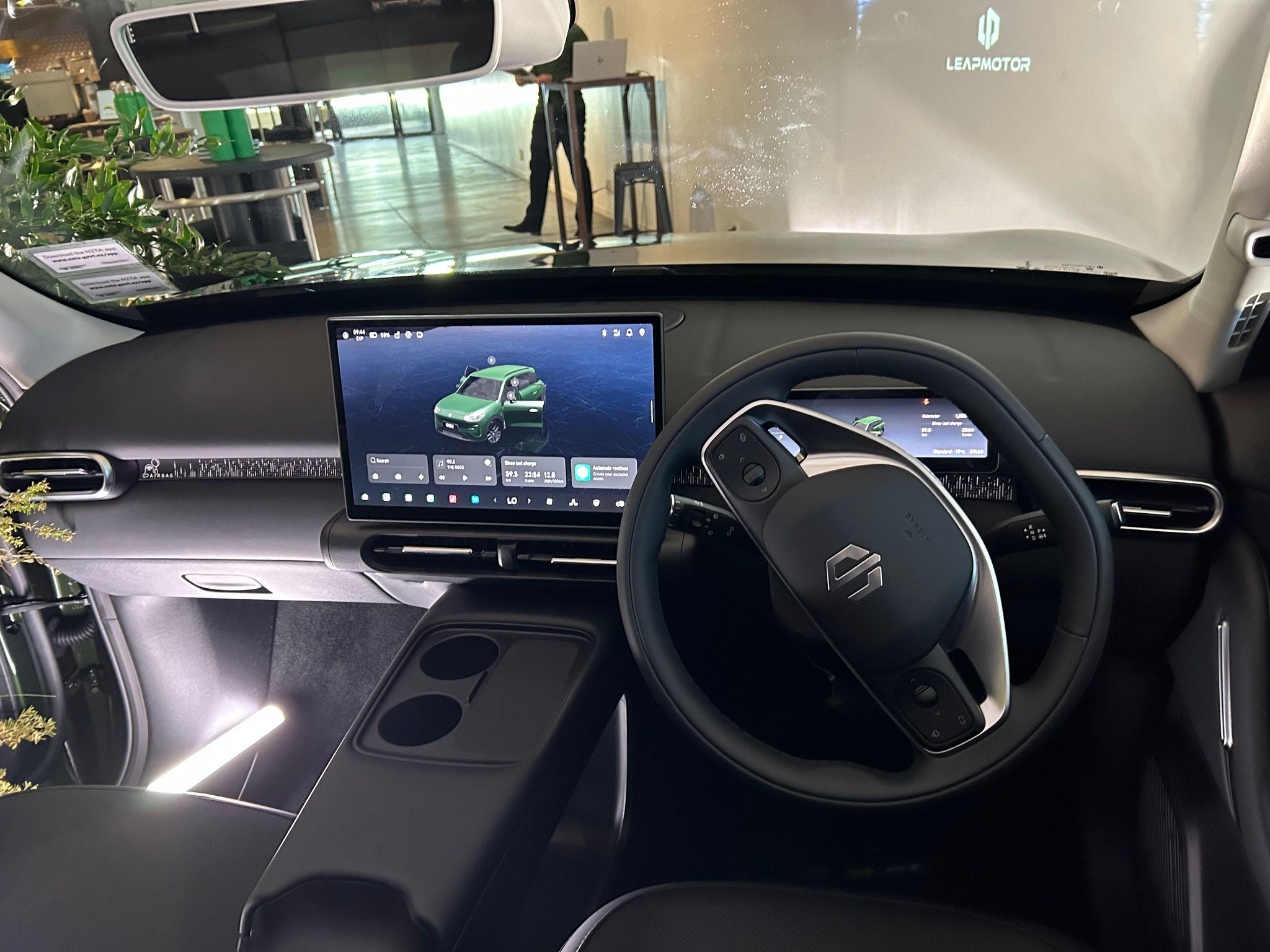 Driver’s-eye view of the Leapmotor B03X interior, showing a right-hand-drive layout with a multifunction steering wheel, digital instrument cluster, and large centrally mounted touchscreen displaying vehicle controls. The minimalist dashboard features slim air vents and textured trim, while a floating centre console includes two cupholders and storage space. The vehicle is displayed indoors at a launch event, with decorative plants, lighting equipment, and Leapmotor branding visible through the windscreen.