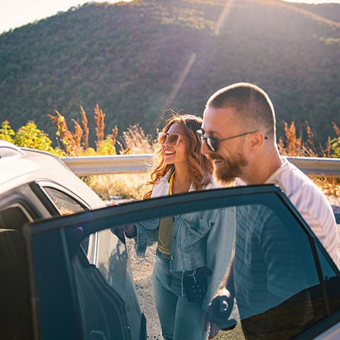 Two people standing beside a white SUV on a roadside overlook, with one person opening the vehicle door. Sunlight illuminates the scene, and tree-covered hills rise in the background, suggesting a scenic road trip or outdoor travel stop.