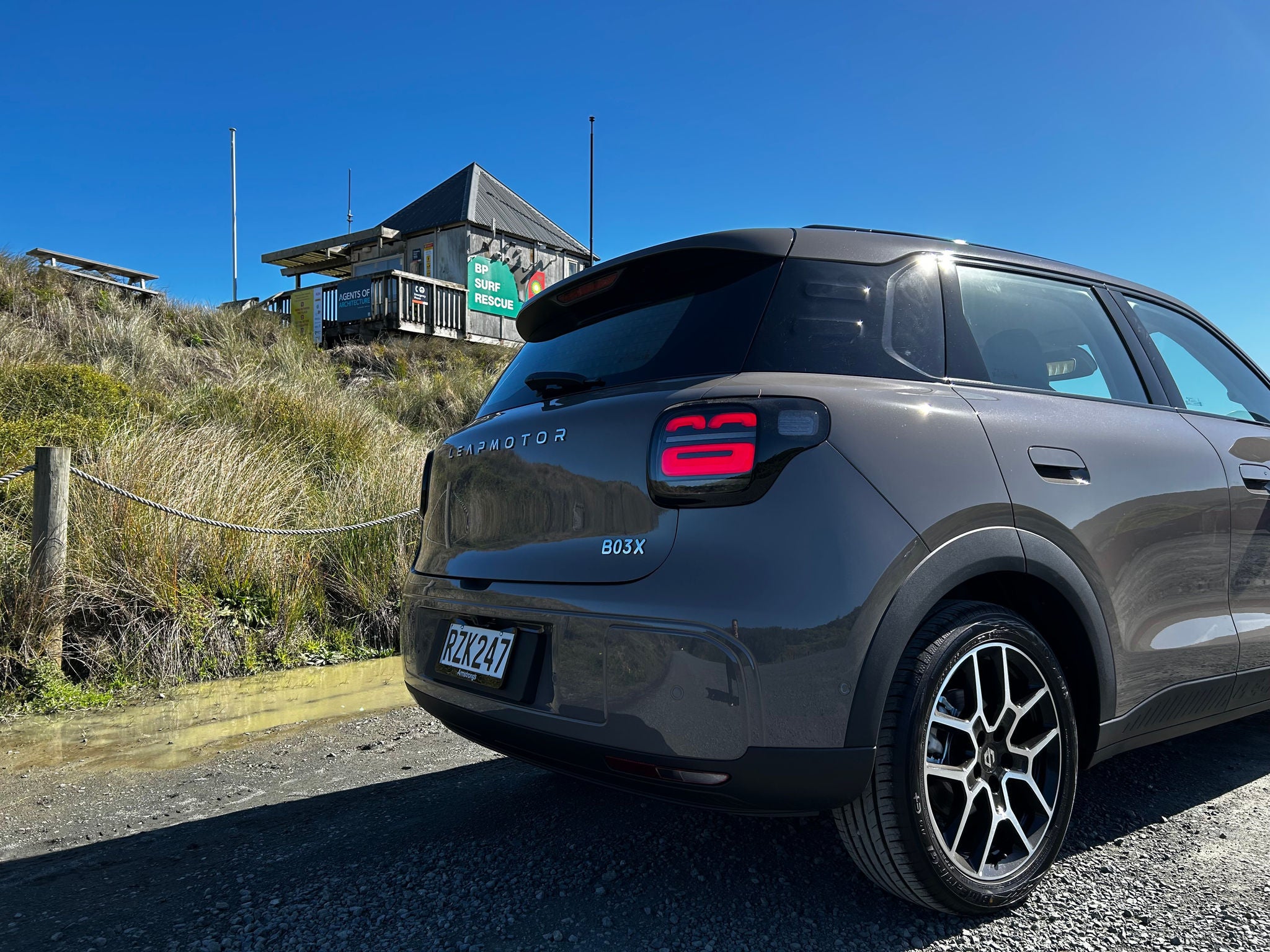 Rear three-quarter view of a light-grey Leapmotor B03X electric SUV parked on a gravel road beside coastal sand dunes. The image highlights the vehicle’s rear design, including illuminated LED taillights, LEAPMOTOR lettering across the tailgate, a B03X badge, and multi-spoke alloy wheels. In the background, a surf rescue building sits atop a grassy dune under a clear blue sky, while bright sunlight reflects off the vehicle’s paintwork and rear window.