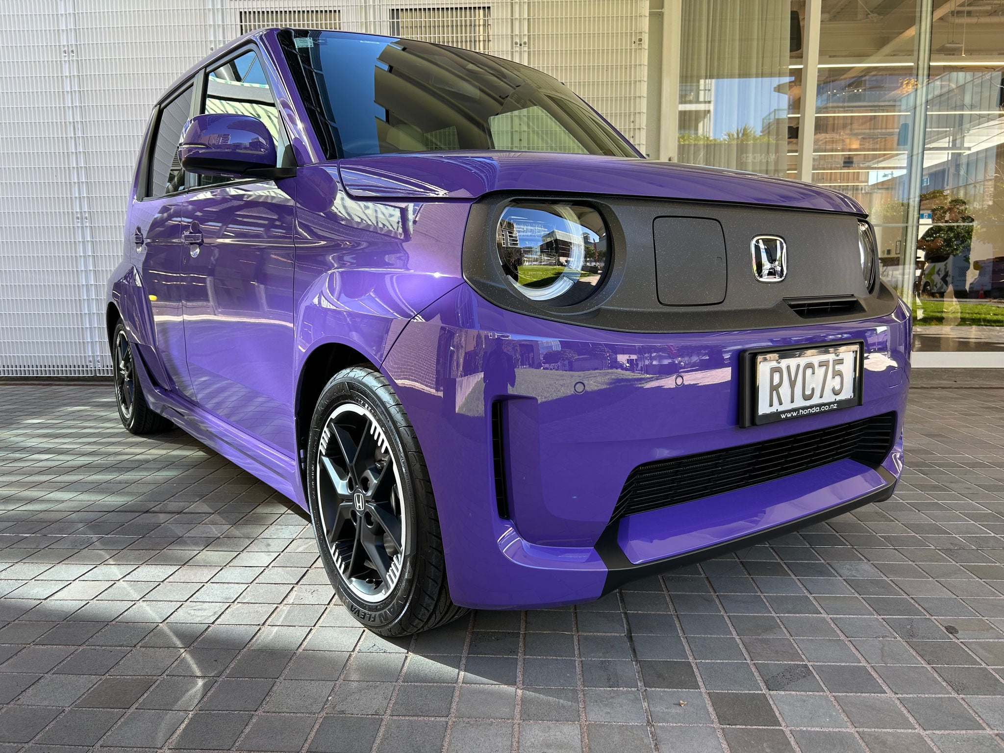 Purple compact Honda electric hatchback parked on a tiled forecourt outside a modern building. The photo is taken from a low front three-quarter angle, showing the car’s boxy design, circular LED headlights, black grille panel with Honda badge, and black alloy wheels. Large glass windows and white architectural panels are reflected in the vehicle’s glossy paint and windshield. A New Zealand license plate reading “RYC75” is visible on the front.