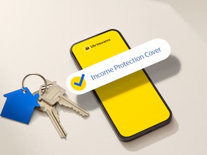 Top-down view of a smartphone displaying a life insurance app on a bright yellow screen with a floating “Income Protection Cover” confirmation badge. A set of house keys with a blue house-shaped keyring lies beside the phone, while stacked cardboard moving boxes sit in the background. Soft natural light casts gentle shadows across the light-colored surface, creating a clean and modern insurance-themed scene focused on homeownership and financial protection.