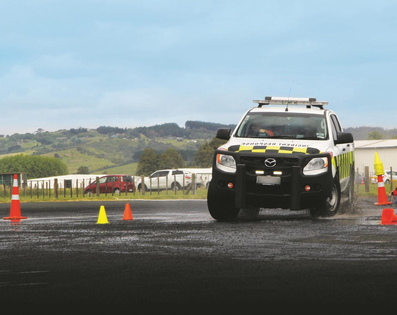 A white incident response pickup truck drives across a wet asphalt surface marked with orange and yellow traffic cones, kicking up water spray. The vehicle has high-visibility yellow and checkered markings, a front bull bar, and a roof-mounted light bar. In the background, there are grassy hills, a fence line, parked cars, and a low building under a cloudy sky.