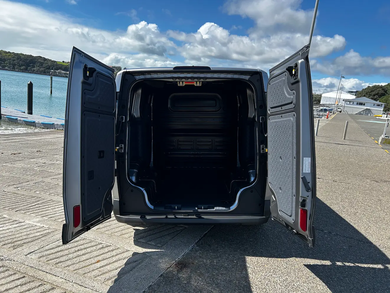 Rear view of a Kia PV5 cargo van with both rear doors open, revealing an empty cargo bay beside a waterfront.