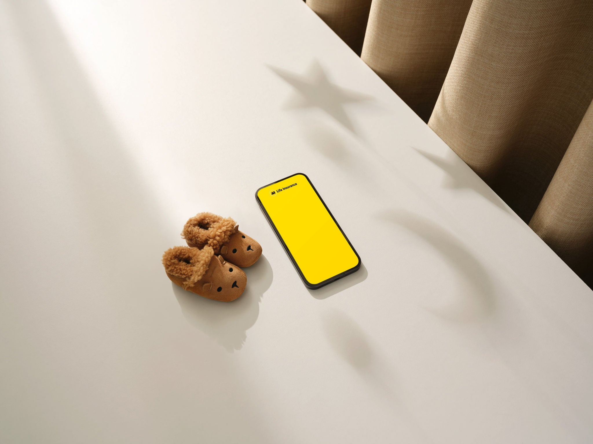 A smartphone displaying a bright yellow life insurance app screen sits on a white tabletop beside a pair of brown fleece-lined children's slippers shaped like animal faces. Warm sunlight streams across the surface, casting soft shadows, including star-shaped silhouettes from an unseen hanging decoration. Beige curtains in the background create a cozy, home-like atmosphere with ample negative space around the objects.