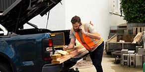 Man in an orange fluoro vest writing on planks of wood that are being loaded into the back of a ute.