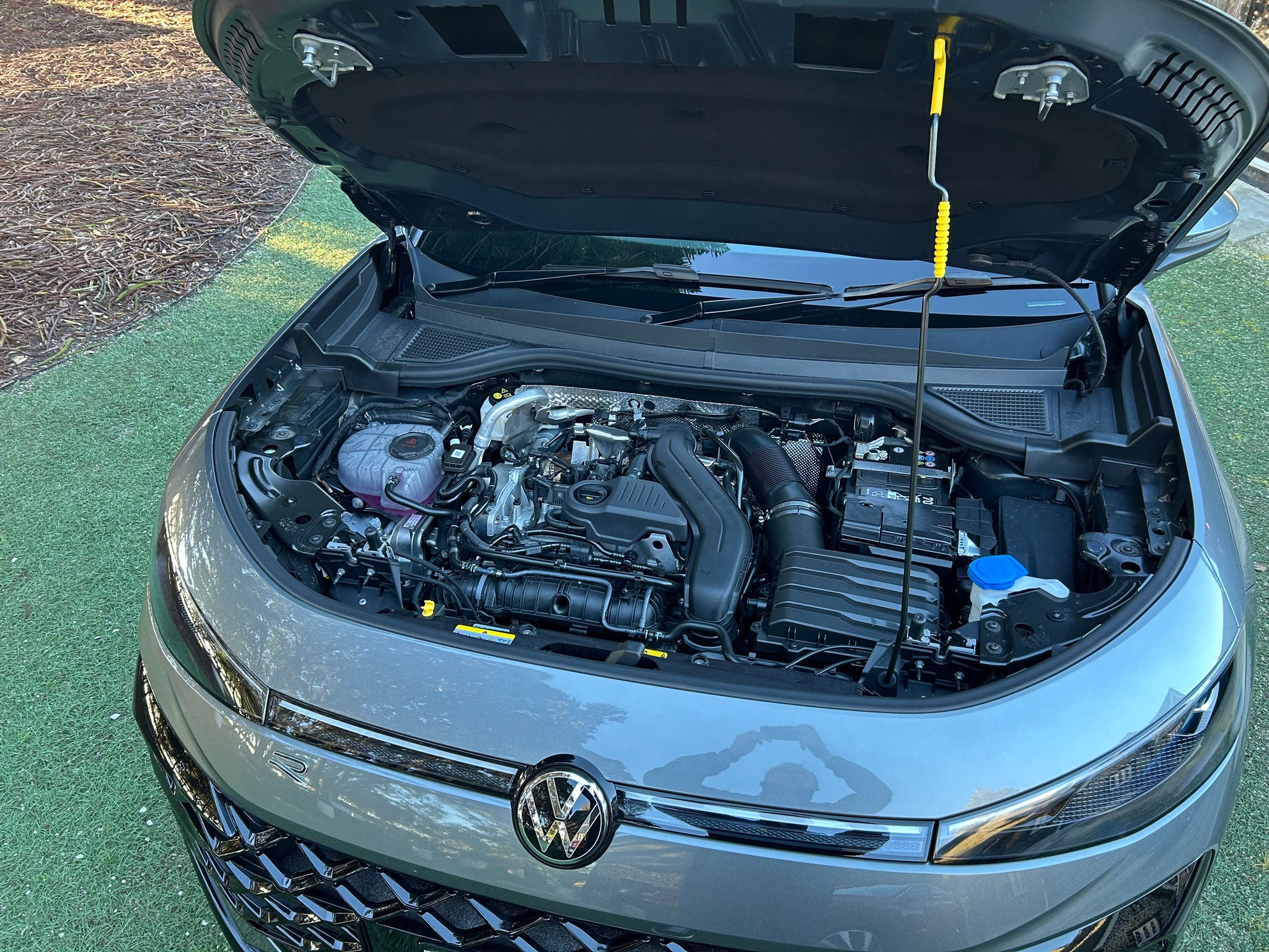 Overhead view of the engine bay of a silver Volkswagen T-Roc R-Line SUV with the bonnet open. Visible components include the engine cover, air intake system, battery, coolant reservoir containing pink fluid, washer fluid filler cap, and various hoses and wiring. The bonnet is supported by a prop rod, and the front Volkswagen badge, slim LED headlights, and upper grille are visible at the bottom of the frame. The vehicle is parked outdoors on a grass-covered surface in daylight.