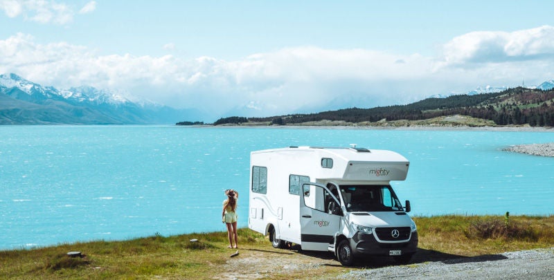 Mighty campervan parked next to scenic South Island lake with young woman standing next to it