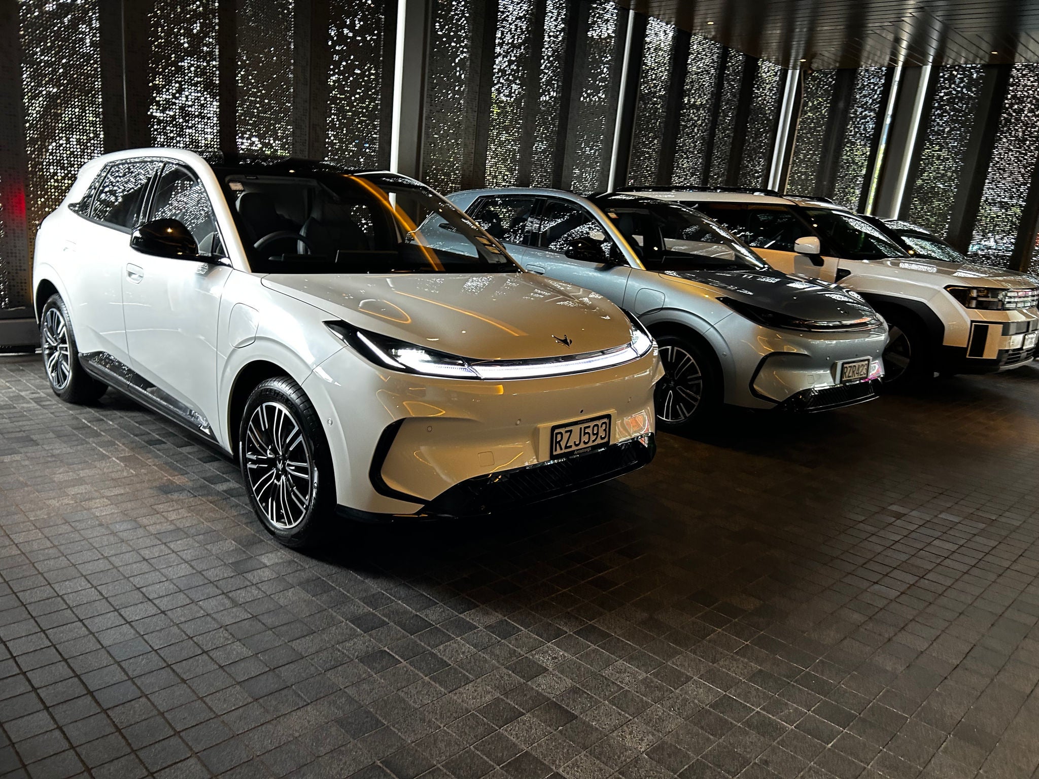 White Arcfox T1 electric SUV displayed indoors beside two other vehicles, viewed from the front-left angle.
