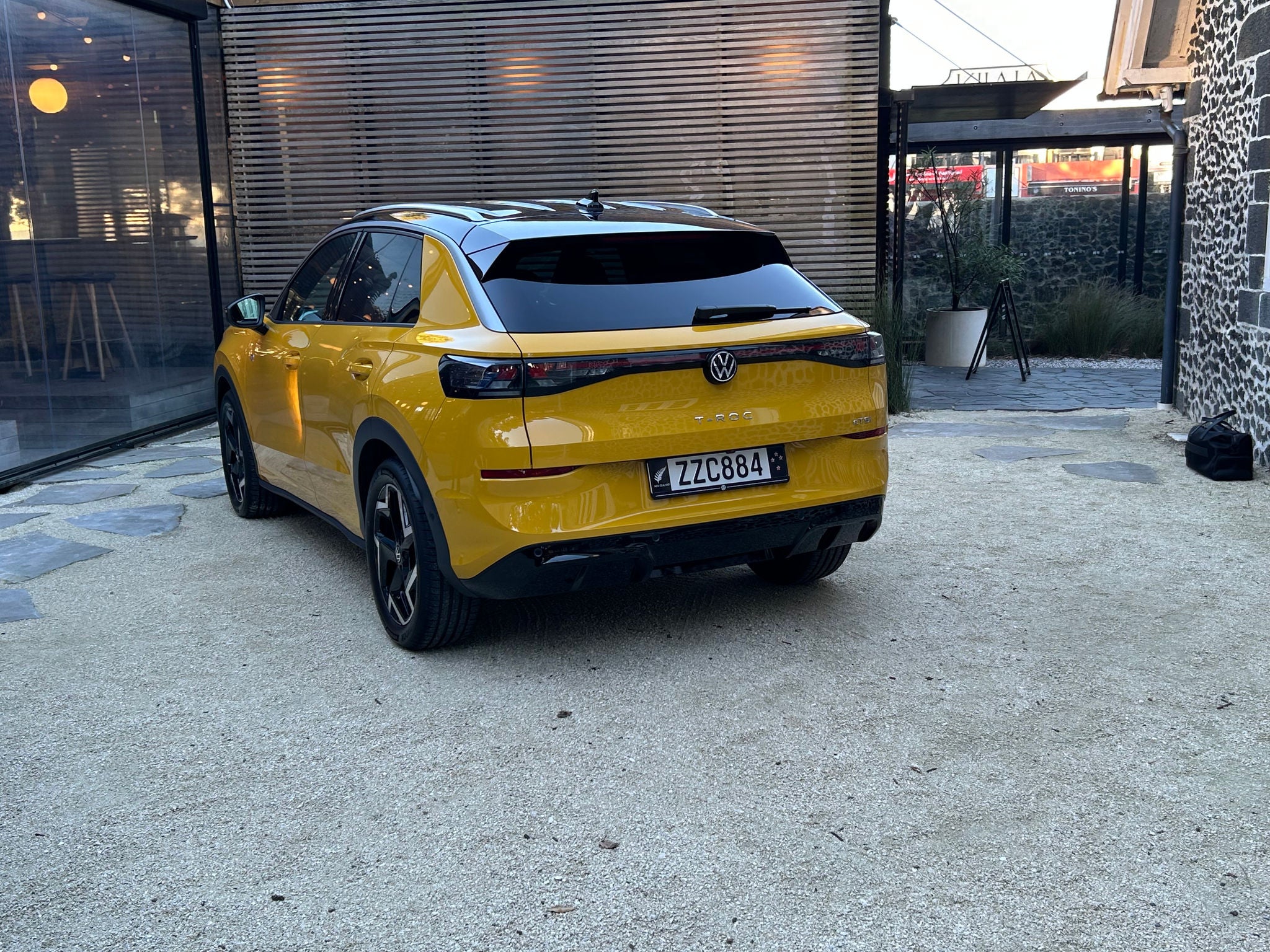 Rear three-quarter view of a yellow Volkswagen T-Roc SUV parked on a light-coloured paved courtyard. The vehicle features a black roof, tinted rear windows, slim LED taillights connected by a dark trim strip, and black alloy wheels. The Volkswagen badge and “T-Roc” model name are visible on the tailgate above a New Zealand licence plate. Surrounding the vehicle are a modern glass-fronted building, timber privacy screening, landscaping elements, and a historic stone building, all illuminated by natural daylight.
