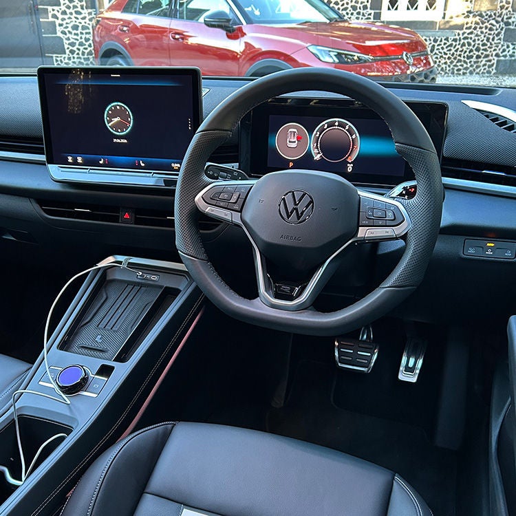 Driver’s-eye view of the interior of a Volkswagen T-Roc R-Line, showing a multifunction steering wheel with the Volkswagen logo, a fully digital instrument cluster, and a central touchscreen infotainment display. The centre console includes a wireless charging pad, rotary control, and storage compartment, while metal sport pedals are visible in the footwell. Black upholstery with contrast stitching and ambient lighting accents enhance the cabin. Through the windscreen, a red Volkswagen SUV and a stone building can be seen parked outside in daylight.