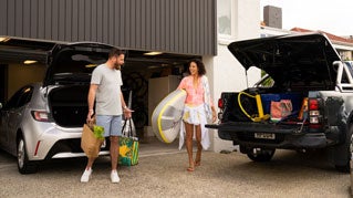 Two people stand in a driveway outside an open garage beside two vehicles with their rear hatches lifted.