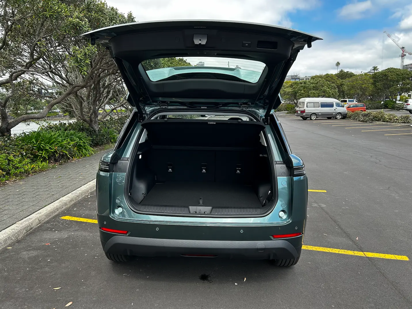 Rear view of a dark green JAECOO J5 SUV with the tailgate open, revealing the luggage compartment in a waterfront car park.