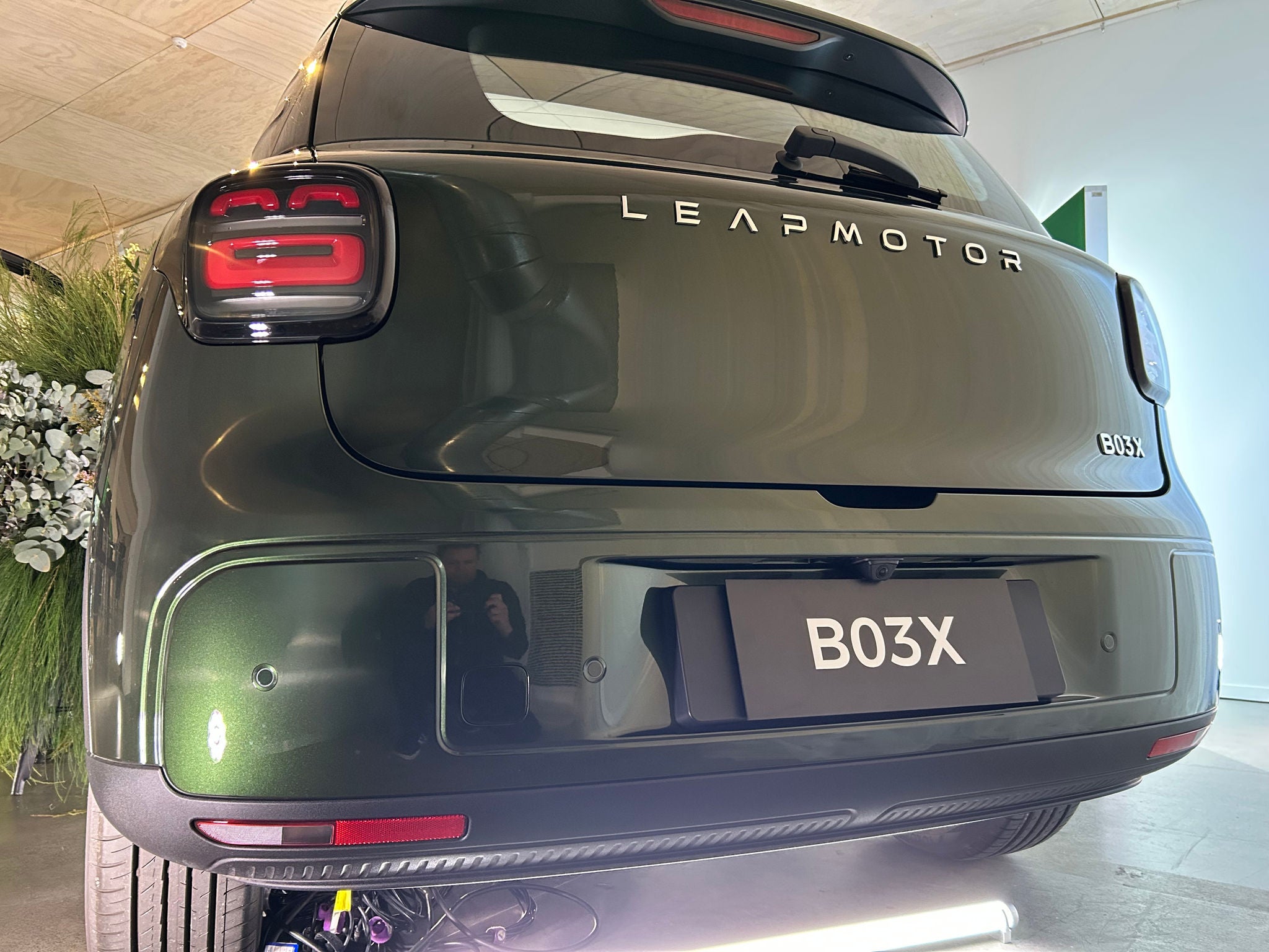 Rear three-quarter view of a dark green Leapmotor B03X compact SUV on display indoors. The vehicle features illuminated rectangular taillights, prominent LEAPMOTOR lettering across the tailgate, and a B03X model badge and display plate. Decorative greenery and white flowers surround the display, while bright showroom lighting reflects off the vehicle’s glossy paint and polished concrete floor.