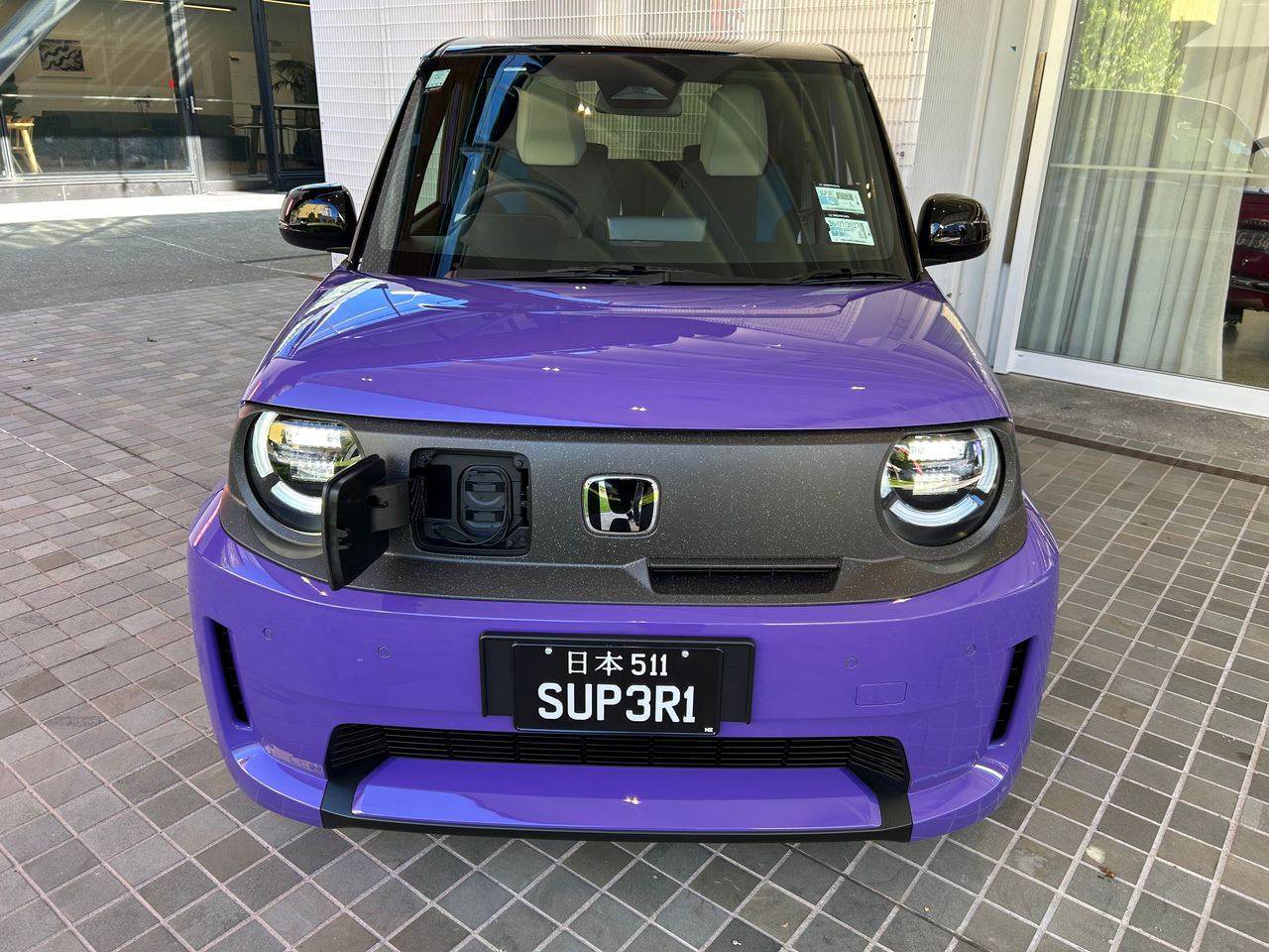 Front view of a purple Honda electric city car parked on a tiled forecourt outside a modern building. The car features a boxy design, black roof, circular LED headlights, and a dark front panel with a chrome Honda badge. The charging port cover is open on the front fascia, revealing the charging socket. A black novelty license plate reading “SUP3R1” with Japanese characters above is mounted on the front bumper. Large windows, white wall panels, and reflected building details are visible in the background, while sunlight creates reflections across the bonnet and windscreen.  Provide your feedback on BizChat