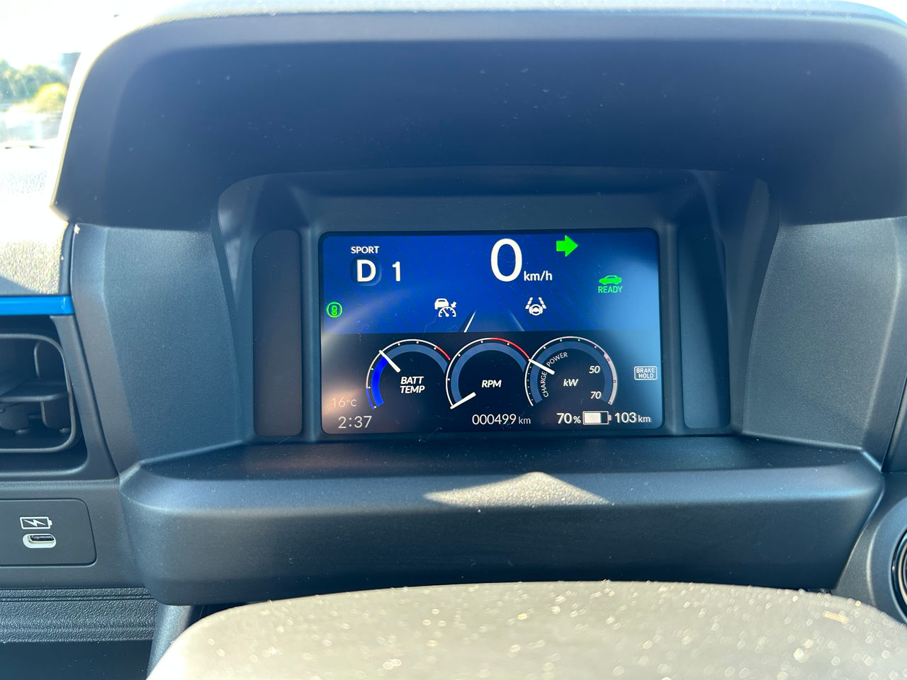 Close-up view of the digital instrument cluster inside a Honda electric city car. The display shows the vehicle stationary at 0 km/h in Sport mode (D1) with a green right-turn indicator illuminated. Three circular gauges display battery temperature, RPM/drive system status, and power/charge output in kW. Additional icons indicate driver assistance features, vehicle readiness, and brake hold. Along the bottom of the screen, the display shows an outside temperature of 16°C, the time 2:37, an odometer reading of 499 km, and a battery charge level of 70% with an estimated driving range of 103 km. The instrument panel is housed within a black dashboard surround, with part of the steering wheel visible in the foreground.