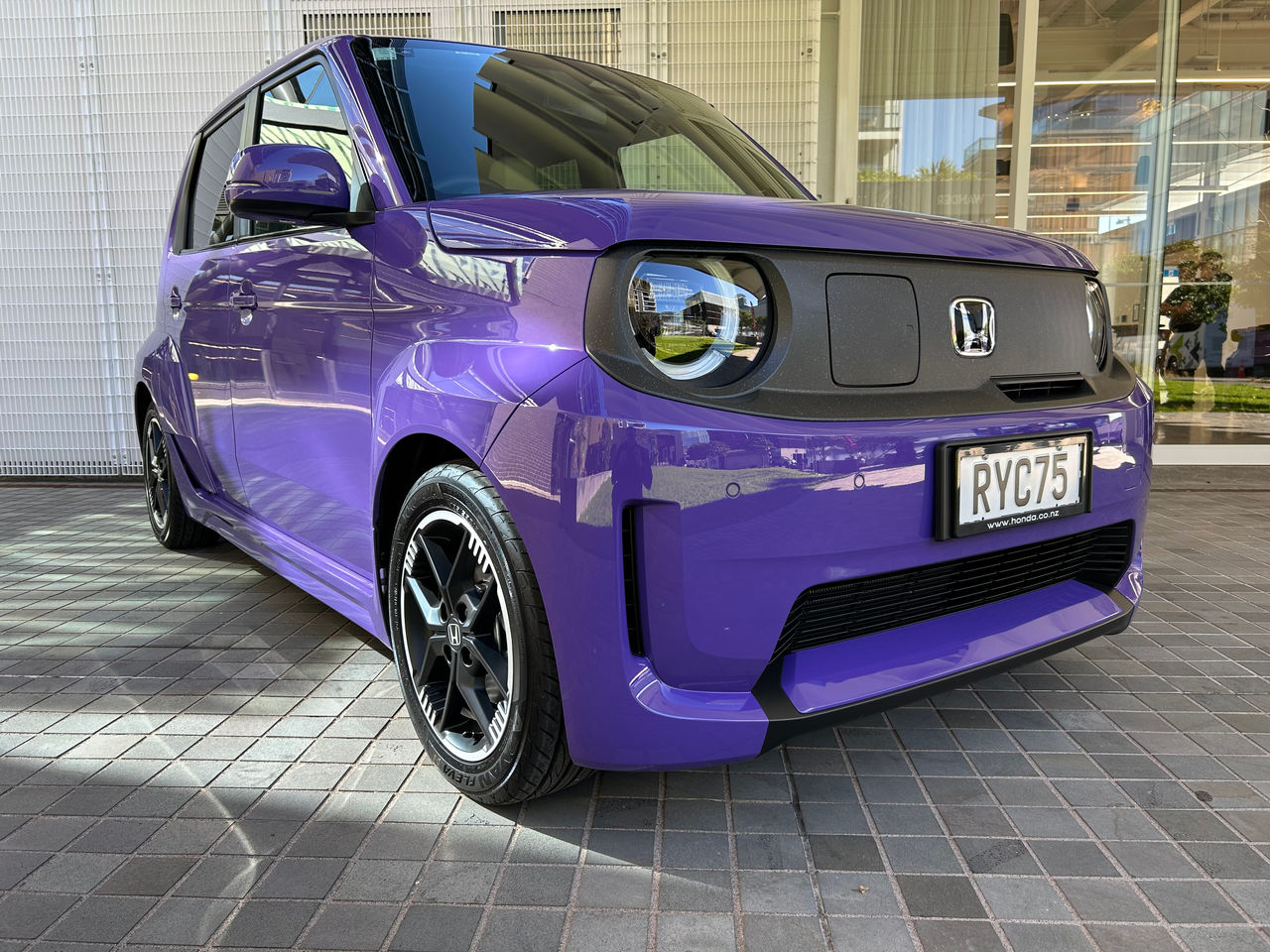 Purple compact Honda electric hatchback parked on a tiled forecourt outside a modern building. The photo is taken from a low front three-quarter angle, showing the car’s boxy design, circular LED headlights, black grille panel with Honda badge, and black alloy wheels. Large glass windows and white architectural panels are reflected in the vehicle’s glossy paint and windshield. A New Zealand license plate reading “RYC75” is visible on the front.