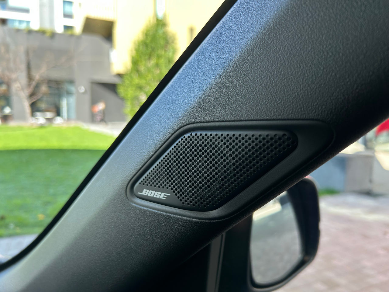 Close-up of a Bose-branded speaker mounted on the front A-pillar inside a Honda electric city car. The black perforated speaker grille is integrated into the textured interior trim beside the windscreen and side window. Part of the exterior side mirror is visible below the speaker, while a blurred background outside the vehicle shows a green lawn, paving, and modern buildings in daylight. The image highlights the vehicle's premium audio system and interior finish.
