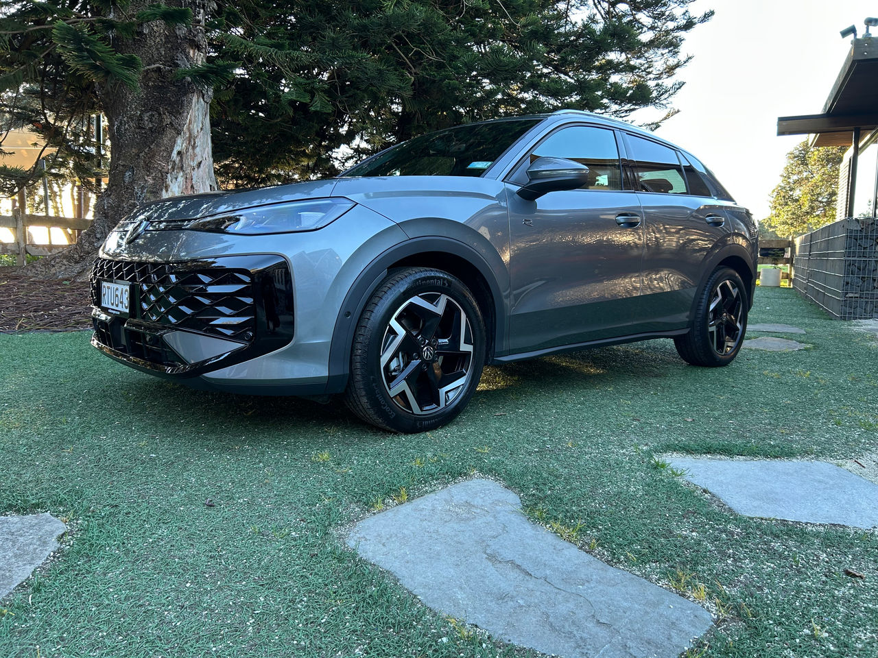 Front three-quarter view of a silver Volkswagen SUV parked on a grass-covered area beside large evergreen trees. The vehicle features a prominent black geometric grille, LED headlights, black alloy wheels, and tinted windows. Stone pavers are visible in the foreground, while a modern building and landscaped garden area appear in the background. The scene is lit by natural daylight, highlighting the SUV's metallic paint finish. 