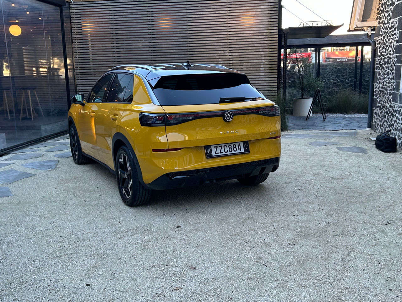 Rear three-quarter view of a yellow Volkswagen T-Roc SUV parked on a light-coloured paved courtyard. The vehicle features a black roof, tinted rear windows, slim LED taillights connected by a dark trim strip, and black alloy wheels. The Volkswagen badge and “T-Roc” model name are visible on the tailgate above a New Zealand licence plate. Surrounding the vehicle are a modern glass-fronted building, timber privacy screening, landscaping elements, and a historic stone building, all illuminated by natural daylight.