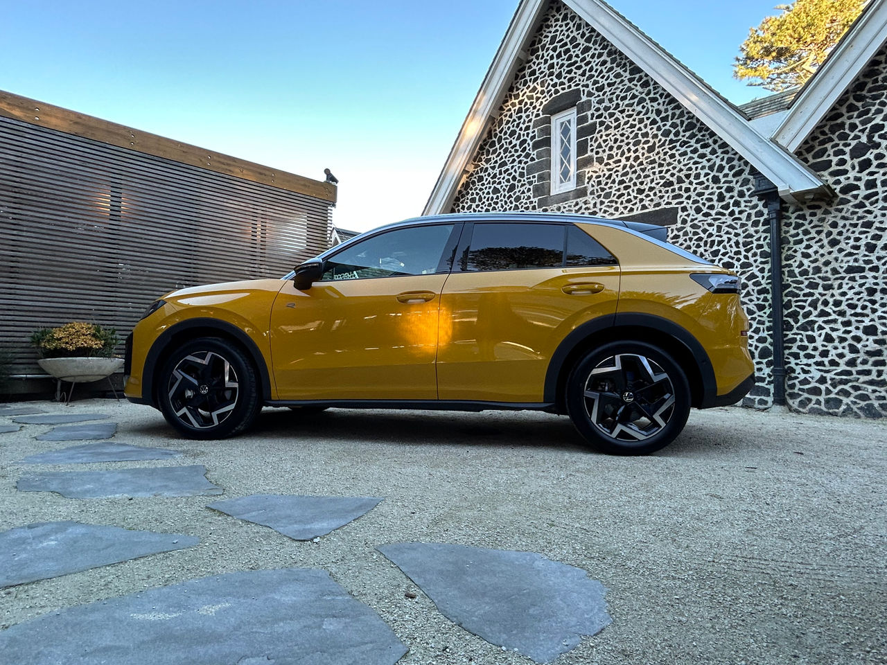 Side profile of a yellow Volkswagen SUV parked on a paved courtyard beside a historic stone building with white-trimmed gables and patterned stone walls. The vehicle features black wheel arch cladding, large two-tone alloy wheels, tinted windows, and a contrasting black roof section. Stone pavers are visible in the foreground, while a timber privacy screen and landscaped planter sit to the left. The scene is illuminated by natural daylight under a clear blue sky. 